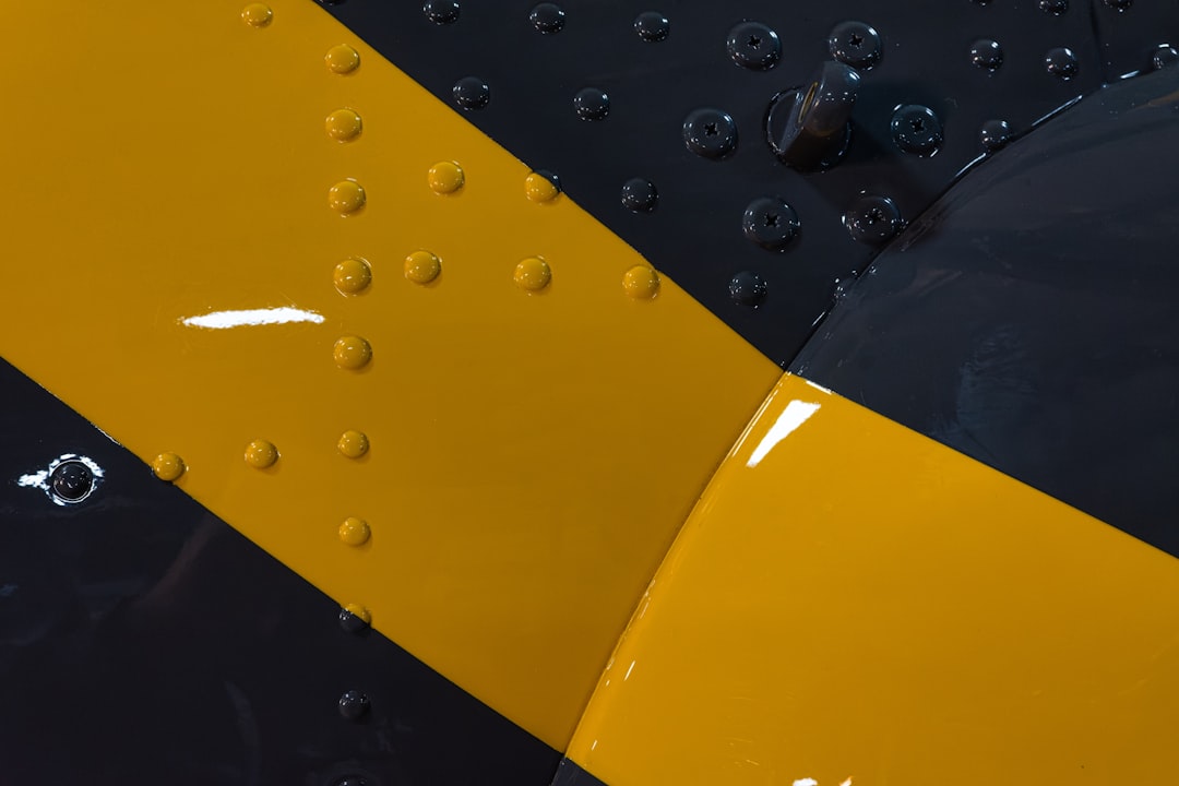 Close Up Of Yellow And Black Painted Airplane Fuselage With Rivets - A close-up view of a yellow and black painted airplane fuselage. The surface of the metal is covered in rivets. There is a section of the plane where the paint is chipped, revealing the metal underneath.